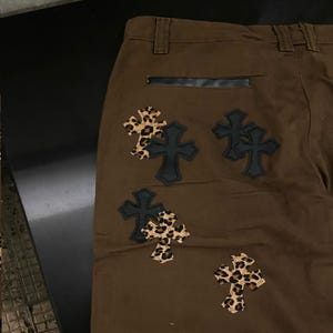 Op de afbeelding: Bruine broek met zwarte leren accenten en kruisvormige patches. De kruisen hebben een luipaardprint en zwarte leren randen. De broek heeft een klassiek ontwerp met vijf zakken.