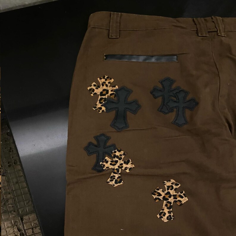 Op de afbeelding: Bruine broek met zwarte leren accenten en kruisvormige patches. De kruisen hebben een luipaardprint en zwarte leren randen. De broek heeft een klassiek ontwerp met vijf zakken.