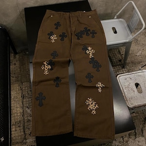 May include: Brown pants featuring a series of scattered cross-shaped patches. The patches alternate between solid black and a tan, black, and brown leopard print pattern. The pants have a standard waistband with a button closure and front pockets.