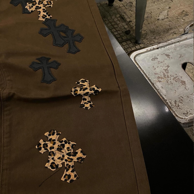 Op de afbeelding: Bruine broek met decoratieve kruisvormige patches. Sommige kruisen zijn zwart, andere hebben een luipaardprint. De broek is gemaakt van een stevige stof.