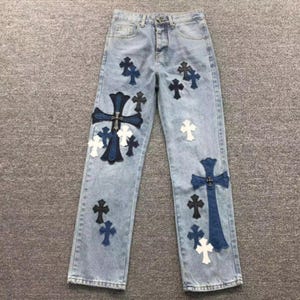 May include: Light blue denim jeans featuring a button fly closure and multiple cross-shaped patches in varying shades of dark blue, black, and white. The patches are distributed across the front of both legs, including two large crosses.