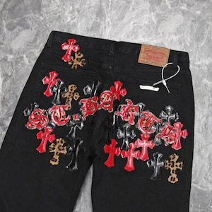 Puede incluir: Vaqueros negros con un dise&ntilde;o de cruces decorativas en rojo, negro y estampado de leopardo. Los vaqueros tienen un dise&ntilde;o cl&aacute;sico de cinco bolsillos y una etiqueta de la marca Levi's.