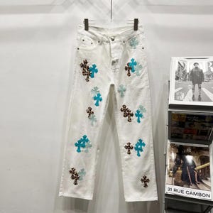 May include: White denim jeans featuring a scattered pattern of cross-shaped patches. The patches alternate between a solid bright turquoise color and a brown leopard print design. The jeans have a standard button fly closure and five pockets.