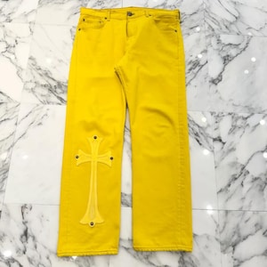 Könnte beinhalten: Helle, gelbe Jeans aus Denim mit einem großen, tonalen, kreuzförmigen Aufnäher am linken unteren Hosenbein. Die Hose verfügt über einen Knopfverschluss, Vordertaschen und Metallnieten, die die dekorative Kreuzapplikation am Stoff befestigen.
