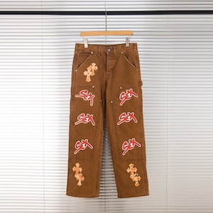 May include: Brown pants featuring a series of red text patches that read "SEX" arranged vertically down each leg, complemented by light-colored cross-shaped patches positioned near the waist and the bottom hem of the garment.
