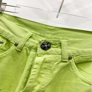 May include: Close-up of bright lime green denim pants. The pants feature a button closure with a decorative silver and black button. The stitching is visible, and the pants are hanging on a clear hanger.