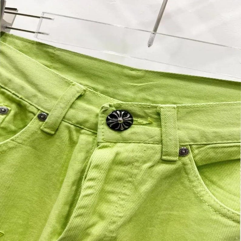 May include: Close-up of bright lime green denim pants. The pants feature a button closure with a decorative silver and black button. The stitching is visible, and the pants are hanging on a clear hanger.