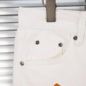 May include: Close-up of white denim jeans with silver rivets on the pocket and waistband. The jeans are hanging on a metal hanger. The pocket has an orange patch at the bottom.