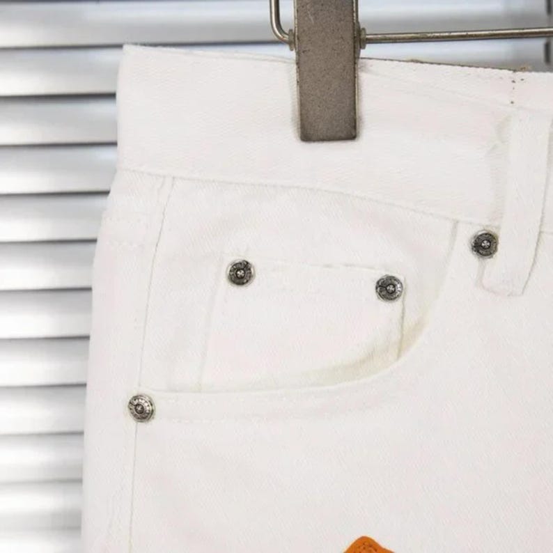 May include: Close-up of white denim jeans with silver rivets on the pocket and waistband. The jeans are hanging on a metal hanger. The pocket has an orange patch at the bottom.