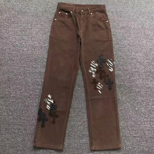 Może przedstawiać: Brązowe dżinsy z dekoracyjnymi aplikacjami w kształcie krzyży w kolorze czarnym, białym i brązowym. Dżinsy mają klasyczny krój z pięcioma kieszeniami oraz zapięcie na guzik i zamek błyskawiczny. Krzyże są rozmieszczone z przodu nogawek.