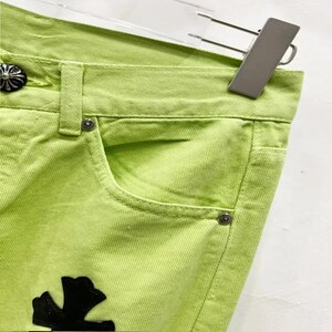 May include: Close-up of lime green denim jeans. The jeans feature a classic five-pocket design with silver-tone rivets and a decorative black cross-shaped patch. The jeans are hanging on a silver hanger.