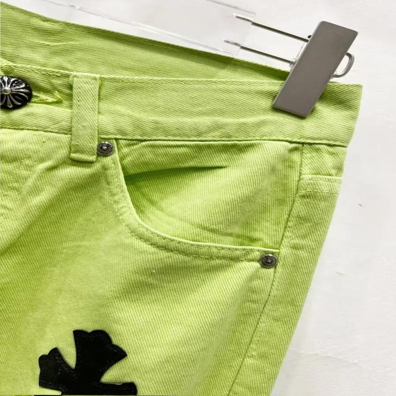 May include: Close-up of lime green denim jeans. The jeans feature a classic five-pocket design with silver-tone rivets and a decorative black cross-shaped patch. The jeans are hanging on a silver hanger.