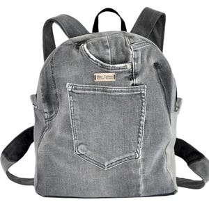 Può includere: Uno zaino in denim grigio con una grande tasca esterna, una maniglia superiore e due spallacci regolabili. Sopra la tasca è cucita una piccola etichetta rettangolare in similpelle con la scritta Mar Calma.