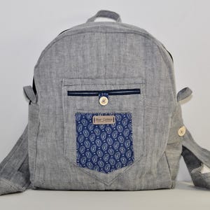 May include: Gray backpack with a front pocket featuring a blue paisley pattern. The backpack has a small top handle, adjustable straps, and a small pocket with a button closure. The brand name "Mar Calmo" is visible on the front pocket.