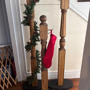 Puede incluir: Tres soportes de madera para calcetines navideños con bases negras. Uno tiene una guirnalda verde con bayas rojas y piñas. Una media acolchada roja cuelga de un soporte. Los soportes están sobre un suelo de madera.