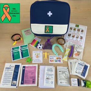 May include: A navy blue first aid kit with a white cross and a variety of items, including bandages, gauze, and a soccer ball. The kit also contains a small notebook, pens, and a rainbow-colored sticker that says "You're Awesome".
