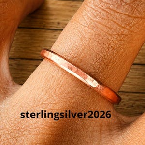 Può includere: Un anello sottile color rame lucido con una fascia liscia e arrotondata. La superficie metallica riflette la luce, mostrando una calda tonalità marrone-arancio. Il testo "sterlingsilver2026" è stampato in lettere nere sotto l'anello nell'immagine.