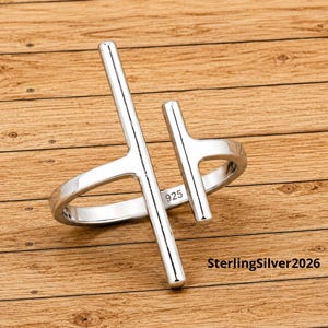 May include: A silver ring with a modern, open design. The ring features two vertical bars connected by a curved band. The text "925" is visible on the ring. The ring is on a wooden surface. The text "SterlingSilver2026" is visible.