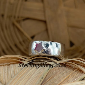 May include: A wide silver band ring featuring a polished metallic finish and a single recessed star shape on the front. The star is filled with a smooth, dark red enamel inlay. The text SterlingSilver2026 is printed in black at the bottom.