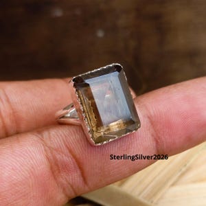 Peut inclure: Bague rectangulaire en quartz fumé avec un anneau en argent. La pierre précieuse est de couleur marron translucide avec une taille à facettes. La bague est posée sur une main, avec le texte "SterlingSilver2026" visible.