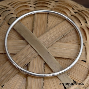 Op de afbeelding: Een glanzende, zilveren armband. De cirkelvormige armband wordt getoond tegen een geweven, lichtbruine achtergrond. De tekst "SterlingSilver2026" is rechtsonder zichtbaar.