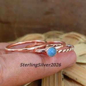 Può includere: Un set di tre anelli color rame. Un anello presenta una fascia sottile e liscia, un altro ha una texture a corda intrecciata e il terzo è una fascia semplice con una piccola pietra cabochon rotonda azzurra. Il testo SterlingSilver2026 è visibile.