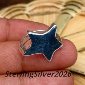 May include: A polished silver ring featuring a prominent five-pointed star design on the front. The metal surface is smooth and reflective, showcasing a clean, solid star shape that sits atop a simple, rounded band. Text reads SterlingSilver2026.