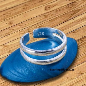 Op de afbeelding: Een zilveren ring met een dubbelbandontwerp, gemarkeerd met "925". De ring wordt gepresenteerd op een blauw, getextureerd oppervlak. De ring is een sieraad.