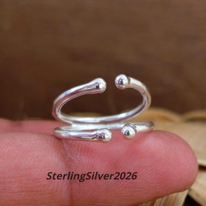 May include: A polished sterling silver ring featuring an open, bypass-style design with two parallel bands. Each end of the bands terminates in a small, rounded ball detail. The text SterlingSilver2026 is visible in the lower portion of the image.