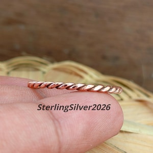 May include: A thin, metallic copper-colored ring featuring a continuous twisted rope design. The surface of the ring has a polished, reflective finish. The text SterlingSilver2026 is printed in black letters across the lower portion of the image.