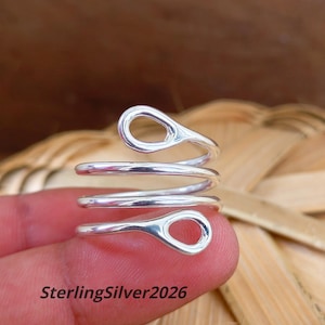 May include: A shiny silver-toned ring featuring a spiral design that wraps around the finger. The ring has two open, rounded ends that taper into loops. The text SterlingSilver2026 is printed in black at the bottom left of the image frame.