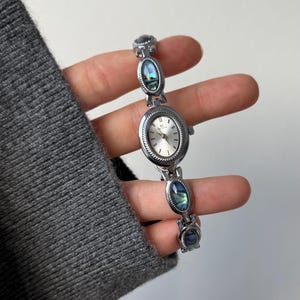 Op de afbeelding: Zilverkleurig polshorloge met een ovale wijzerplaat en een zilveren band. De band heeft drie decoratieve abalone schelp accenten. De wijzerplaat heeft een zilveren wijzerplaat met zwarte uurmarkeringen en wijzers.