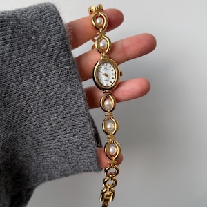 Può includere: Orologio da polso color oro con quadrante ovale bianco e cinturino a maglie dorate. Il cinturino presenta maglie ovali con perle finte incastonate. Il quadrante dell'orologio ha lancette e indici dorati. L'orologio è al quarzo.