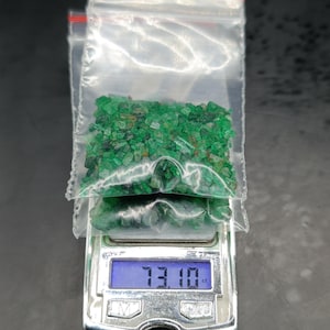 May include: A collection of small, raw green emerald gemstone fragments contained within a clear plastic zip-top bag. The bag rests on a digital scale displaying a weight of 73.10 ct, indicating the total carat weight of the loose green stones.