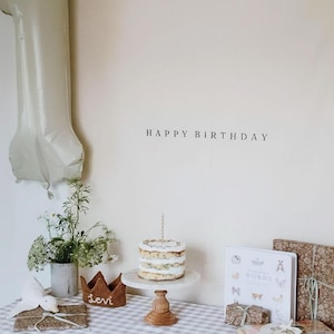 May include: A birthday scene with a small cake on a marble stand, a number one balloon, and the words "HAPPY BIRTHDAY" on the wall. A crown and gifts are on a checkered tablecloth. A vase of flowers is also present.
