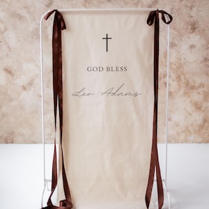 Pode incluir: Uma faixa creme com as palavras "GOD BLESS" e "Leo Adams" em escrita preta, encimada por uma cruz preta. A faixa está suspensa de uma estrutura de metal branco e adornada com fitas marrons.
