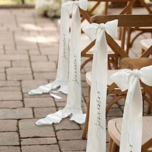 May include: Wooden chairs adorned with white satin ribbons tied in bows, each bearing the word "reserved" in elegant script. The ribbons cascade downwards, enhancing the wedding or event's decor. The chairs are set on a brick patio.