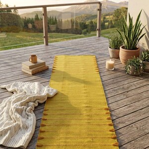 Puede incluir: Una alfombra rectangular amarilla mostaza con l&iacute;neas horizontales y borlas est&aacute; colocada sobre una terraza de madera. Tambi&eacute;n hay una manta de punto de cable crema, libros y plantas en macetas. El fondo muestra una vista panor&aacute;mica de monta&ntilde;as y &aacute;rboles.