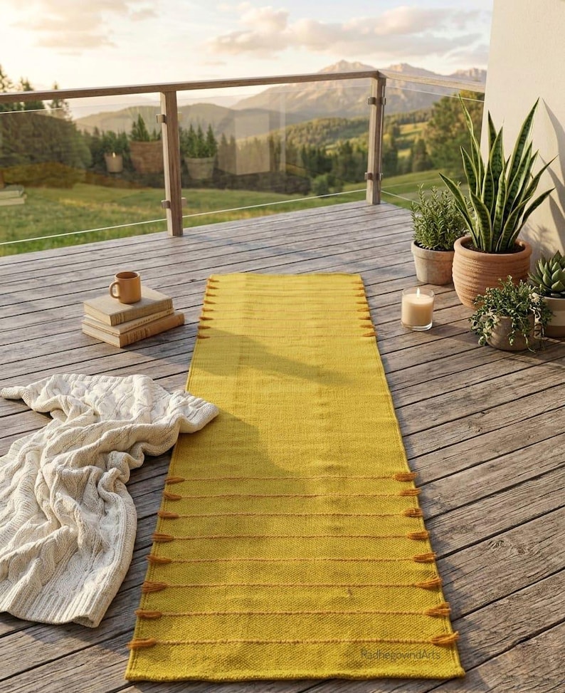 Puede incluir: Una alfombra rectangular amarilla mostaza con l&iacute;neas horizontales y borlas est&aacute; colocada sobre una terraza de madera. Tambi&eacute;n hay una manta de punto de cable crema, libros y plantas en macetas. El fondo muestra una vista panor&aacute;mica de monta&ntilde;as y &aacute;rboles.