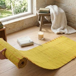 Puede incluir: Una esterilla de yoga amarilla, parcialmente desenrollada, est&aacute; sobre un suelo de madera. Una peque&ntilde;a vela, una toalla blanca doblada y una planta en maceta est&aacute;n cerca. La esterilla mide unos 183 cm de largo.