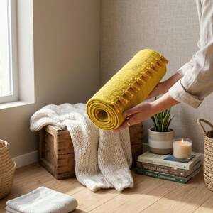 Puede incluir: Una alfombra amarilla enrollada con borlas se muestra. Una manta blanca de punto est&aacute; sobre una caja de madera. Una toalla gris doblada est&aacute; en el suelo. Una vela y libros est&aacute;n cerca.