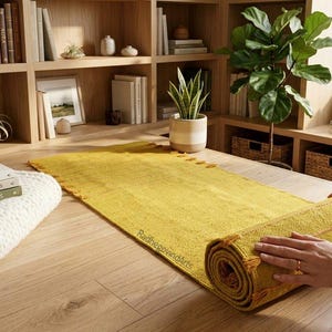 Puede incluir: Una alfombra de pasillo tejida de color amarillo mostaza se enrolla sobre una mesa de madera clara. La alfombra tiene una superficie texturizada y borlas decorativas. Una pila de libros, una vela y una manta blanca est&aacute;n sobre la mesa.