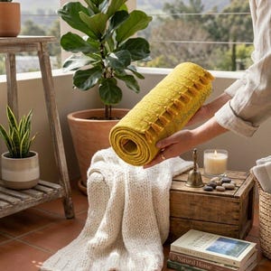 Puede incluir: Una manta amarilla enrollada con flecos es sostenida. Una manta de punto blanca est&aacute; sobre una caja de madera. La escena incluye plantas en macetas, libros y una vela, sugiriendo un ambiente exterior acogedor.
