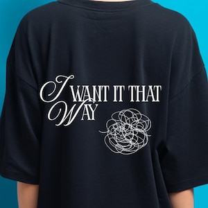 Puede incluir: Una camiseta negra con la frase "I WANT IT THAT WAY" impresa en escritura blanca y un diseño de garabatos. La persona está de espaldas a la cámara sobre un fondo azul.