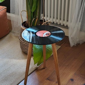 May include: A three-legged side table featuring a circular top made from a black vinyl record. The record has a central red label with black text and a white graphic design. The table has three slender, light-toned wooden legs.