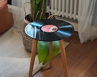 May include: A three-legged side table featuring a circular top made from a black vinyl record. The record has a central red label with black text and a white graphic design. The table has three slender, light-toned wooden legs.