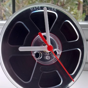 May include: A clock designed as a film reel, with a clear plastic casing and black film inside. The clock features white hands and a red second hand. The words "MADE IN U.S.A." are printed on the top of the clock.