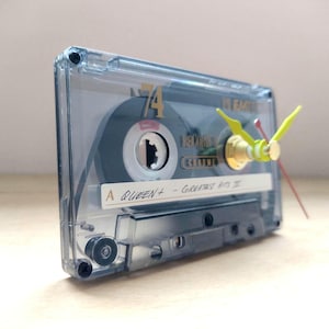 May include: A transparent cassette tape clock with yellow clock hands and a red second hand. The cassette tape features the text "A QUEEN+ - Greatest Hits II". A unique decorative clock.
