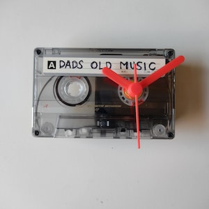 May include: A clear cassette tape transformed into a clock. The clock hands are a vibrant red. The tape label displays "A DADS OLD MUSIC". The clock is fixed to a white wall. A distinctive, retro-style home decoration.