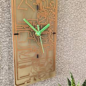 May include: A rectangular wall clock featuring a translucent circuit board base with visible copper traces. The clock has three bright lime green hands centred on the board, with small black markers at the three, six, nine, and twelve o'clock positions.